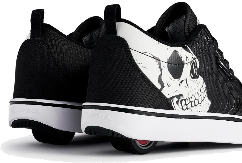 A close-up of Heelys Pro 20 PRNT Glow Boys Shoes in black and white, featuring a glow-in-the-dark skull graphic on the side and low-profile wheels in the heels.