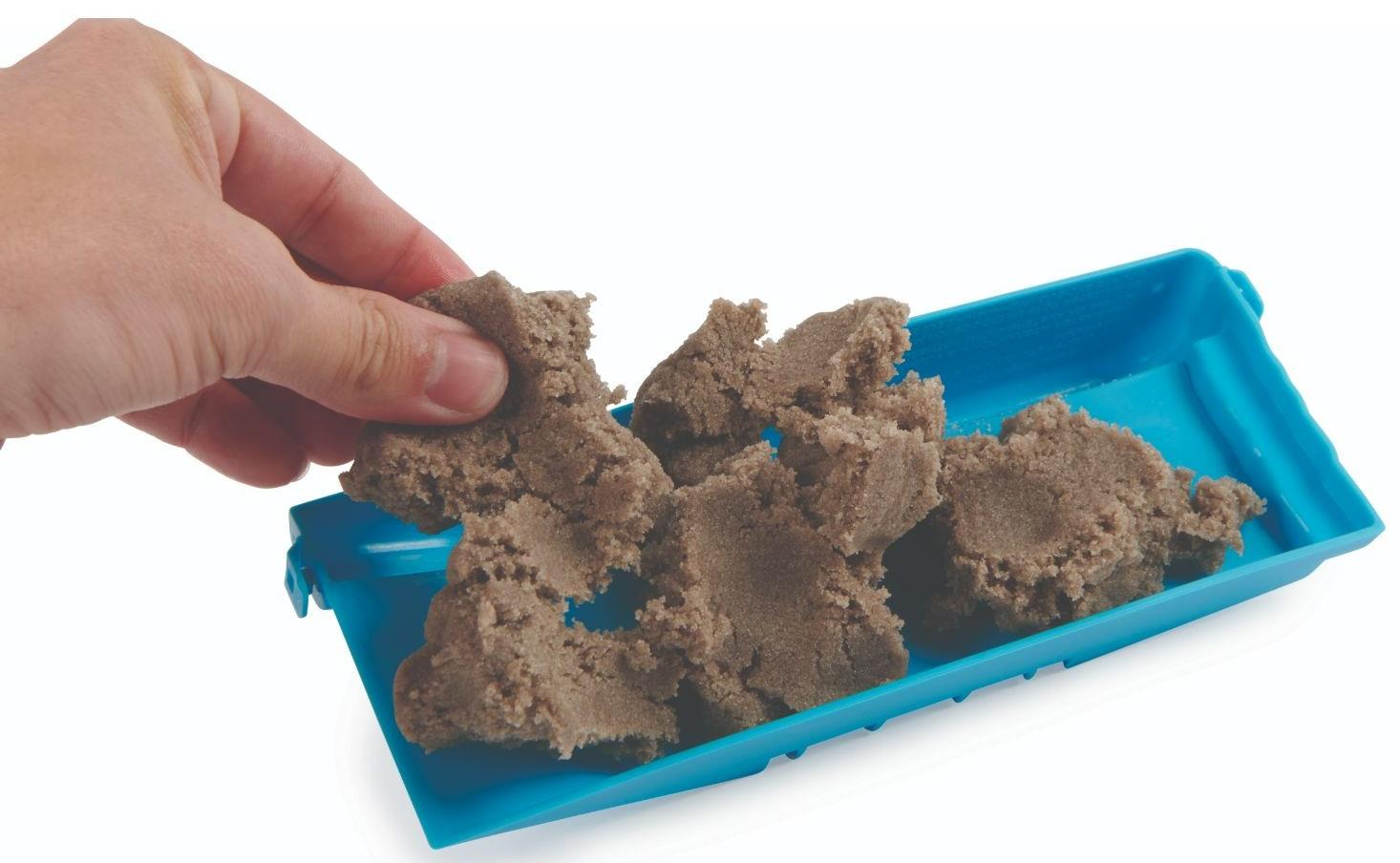 A hand holds brown reusable concrete from the Tech Deck DIY Concrete Reusable Fingerboard Ramp Modelling Set, with extra sand in a blue tray by Tech Deck—ideal for customizing your own fingerboard ramps.
