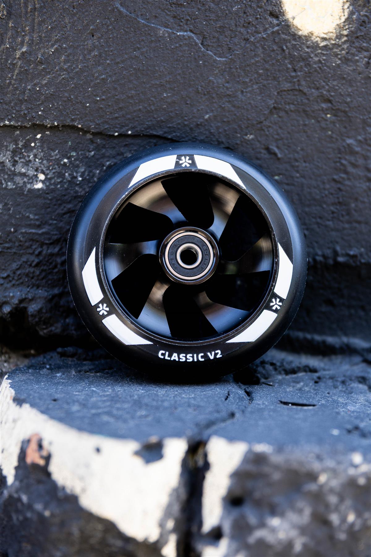 A Union Classic V2 110mm Stunt Scooter Wheel in black with a visible metal bearing leans against a rough, chipped black-and-white painted stone surface.