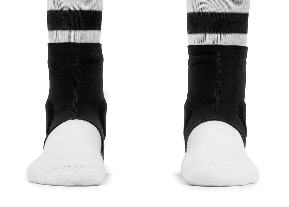 Front view of two feet in white socks with black stripes, wearing REKD Energy Slim Footies Ankle Protection Pad - Black by REKD, covering the ankle and heel for impact protection while leaving toes and soles exposed.