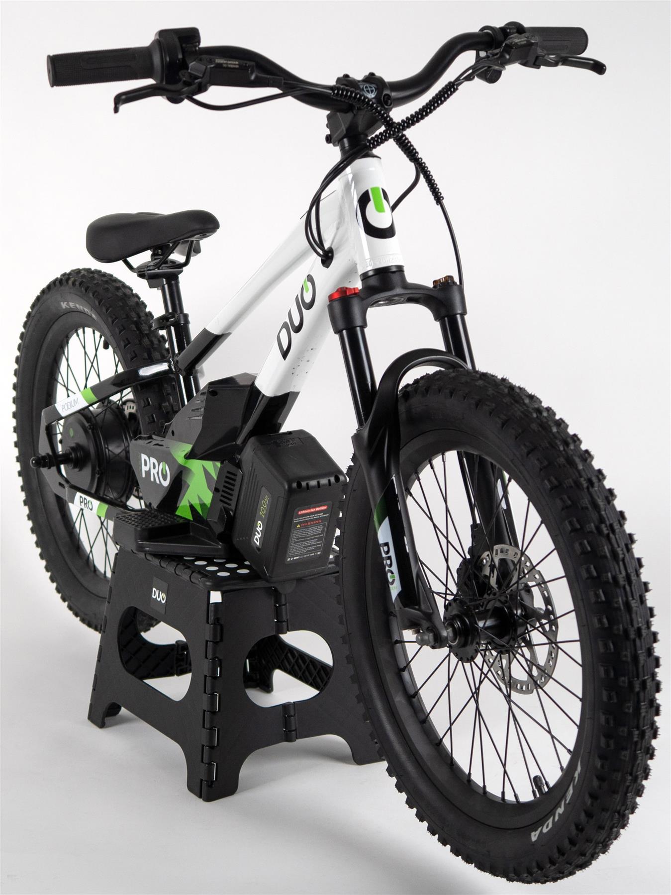 The Duo 20" Podium Pro Kids Electric Balance Bike by Duo, in white and black, features thick tires, green accents, and a visible battery pack. Displayed on a black stand with “DUO” on the frame, it’s perfect for kids seeking an electric balance bike.
