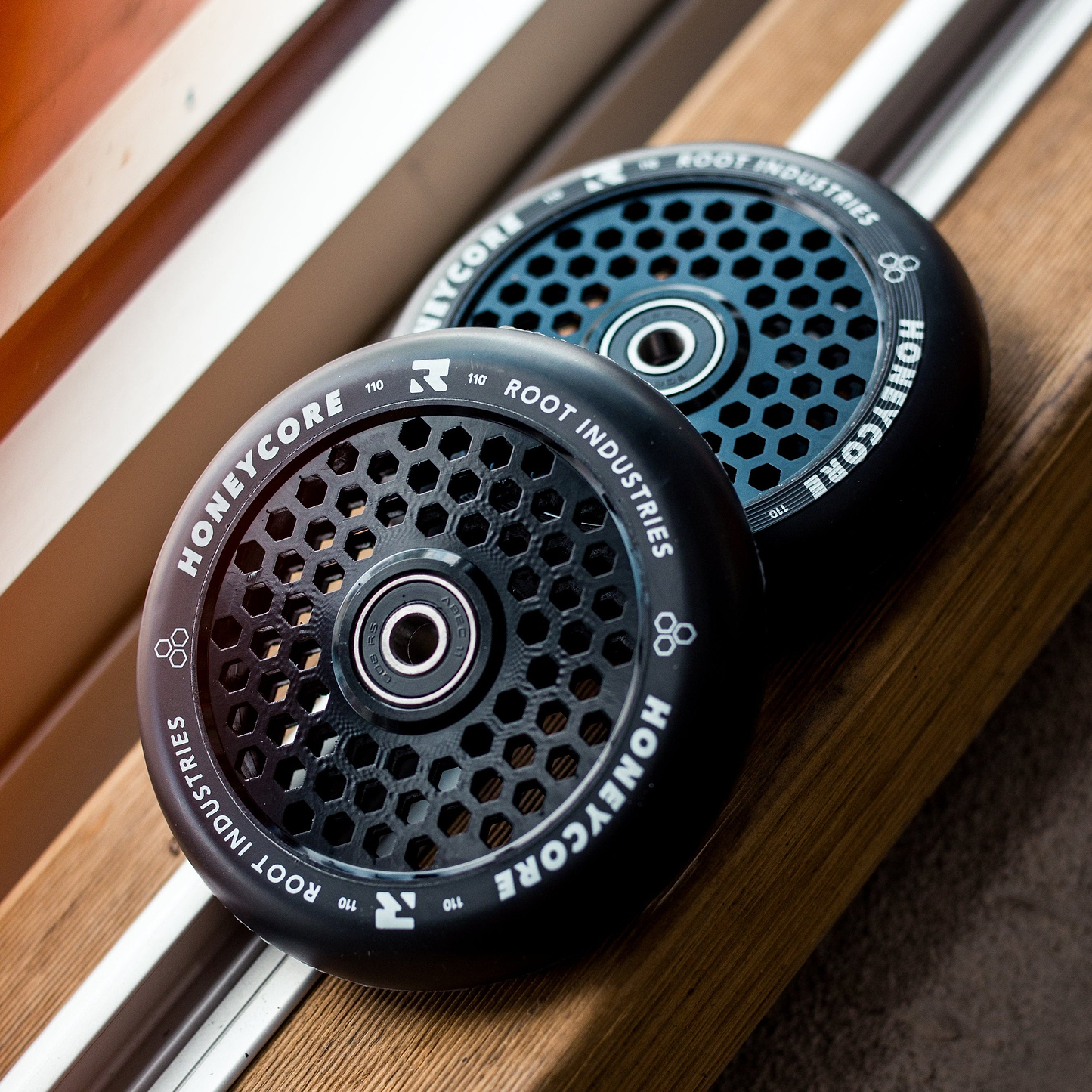 Two Root Industries Honeycore 110mm Stunt Scooter Wheels in Black, featuring the signature honeycomb design, sit side by side on a wooden windowsill.