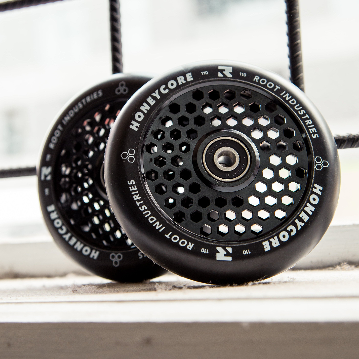Two Root Industries Honeycore 110mm Stunt Scooter Wheels in black, featuring hexagonal cutouts and distinctive branding, rest on a white windowsill with a metal grid and cityscape blurred in the background.