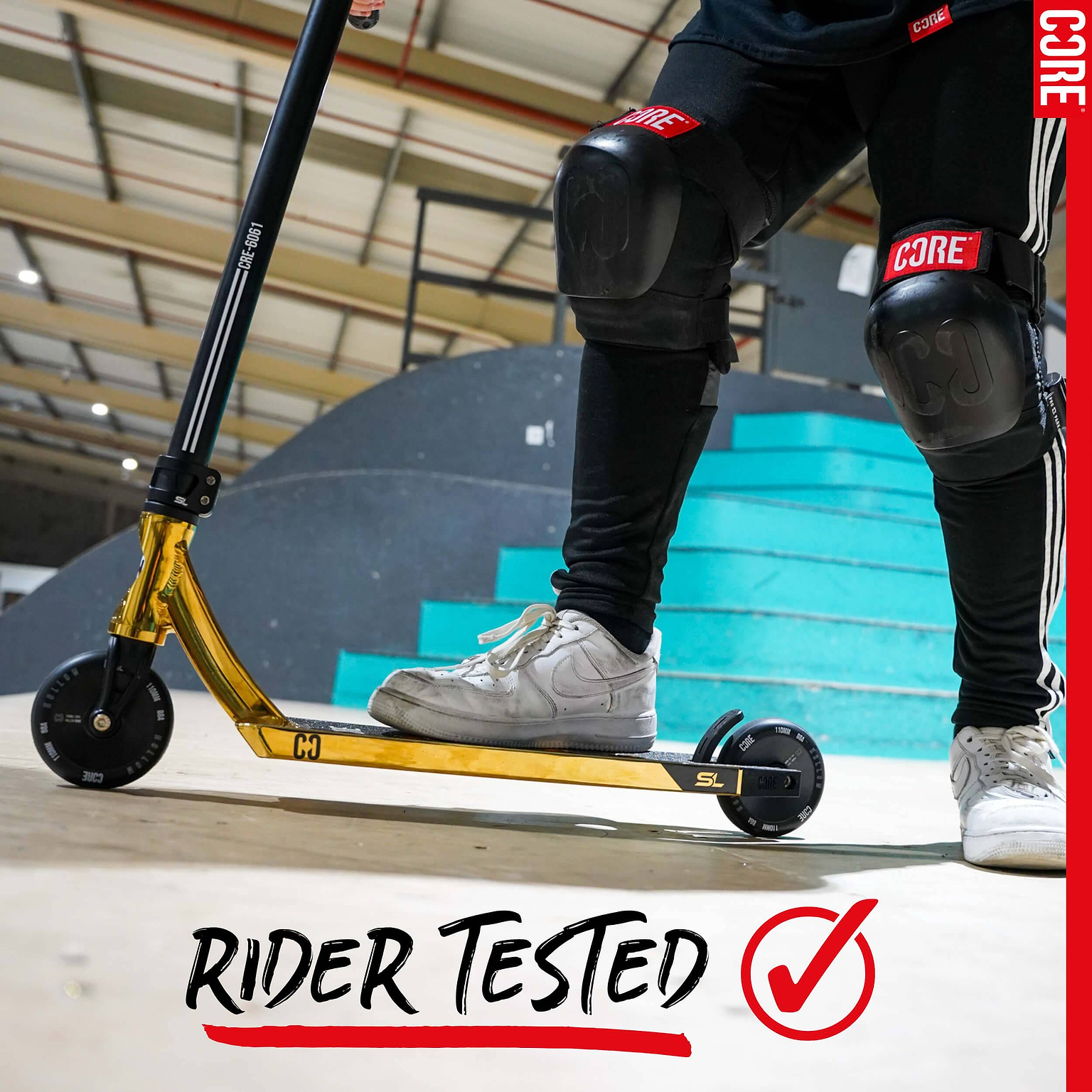 Person wearing black knee pads and white sneakers stands on a gold scooter with CORE Hollow Core V2 100mm Stunt Scooter Wheel - Black at a skate park. Blue stairs, curved ramp, and “RIDER TESTED” text with red checkmark behind them.