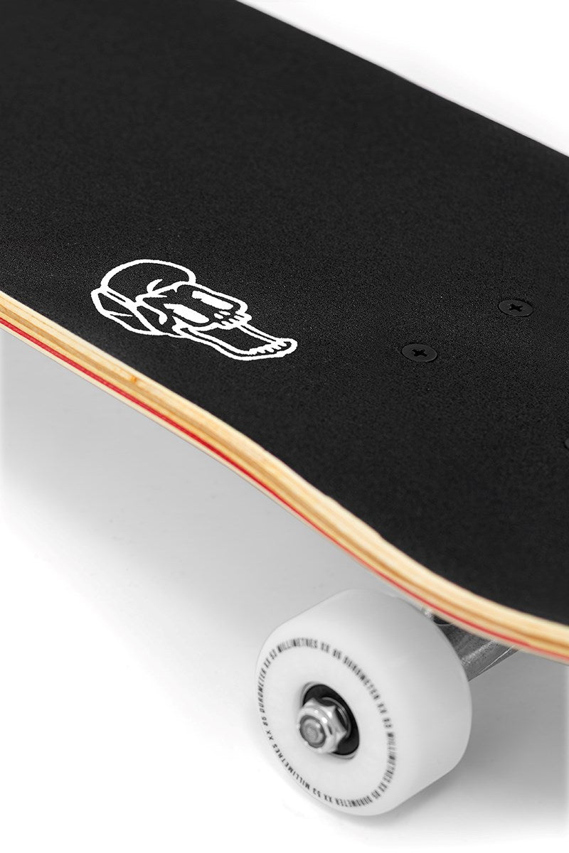 A close-up of the Enuff Meltworld Mini Confused Complete Skateboard (7.25" x 29.5") shows black grip tape with a white skull, a layered wooden edge, and one white wheel with a visible metal axle and bearing.