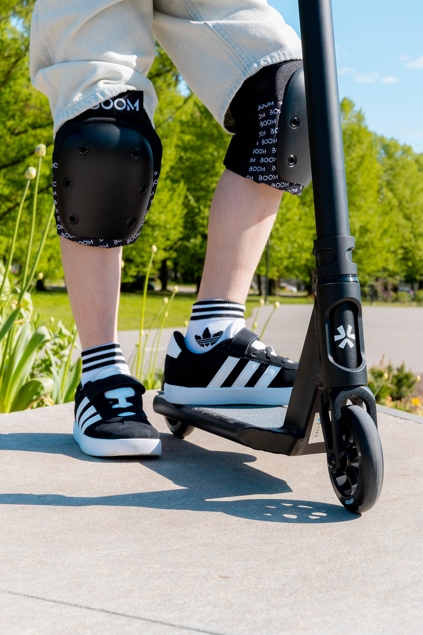 A person wearing knee pads, Adidas sneakers, and striped socks stands outdoors on a sunny day with green trees in the background, riding the Union Aero Complete Stunt Scooter - Black by Union—a perfect lightweight stunt scooter for new riders.