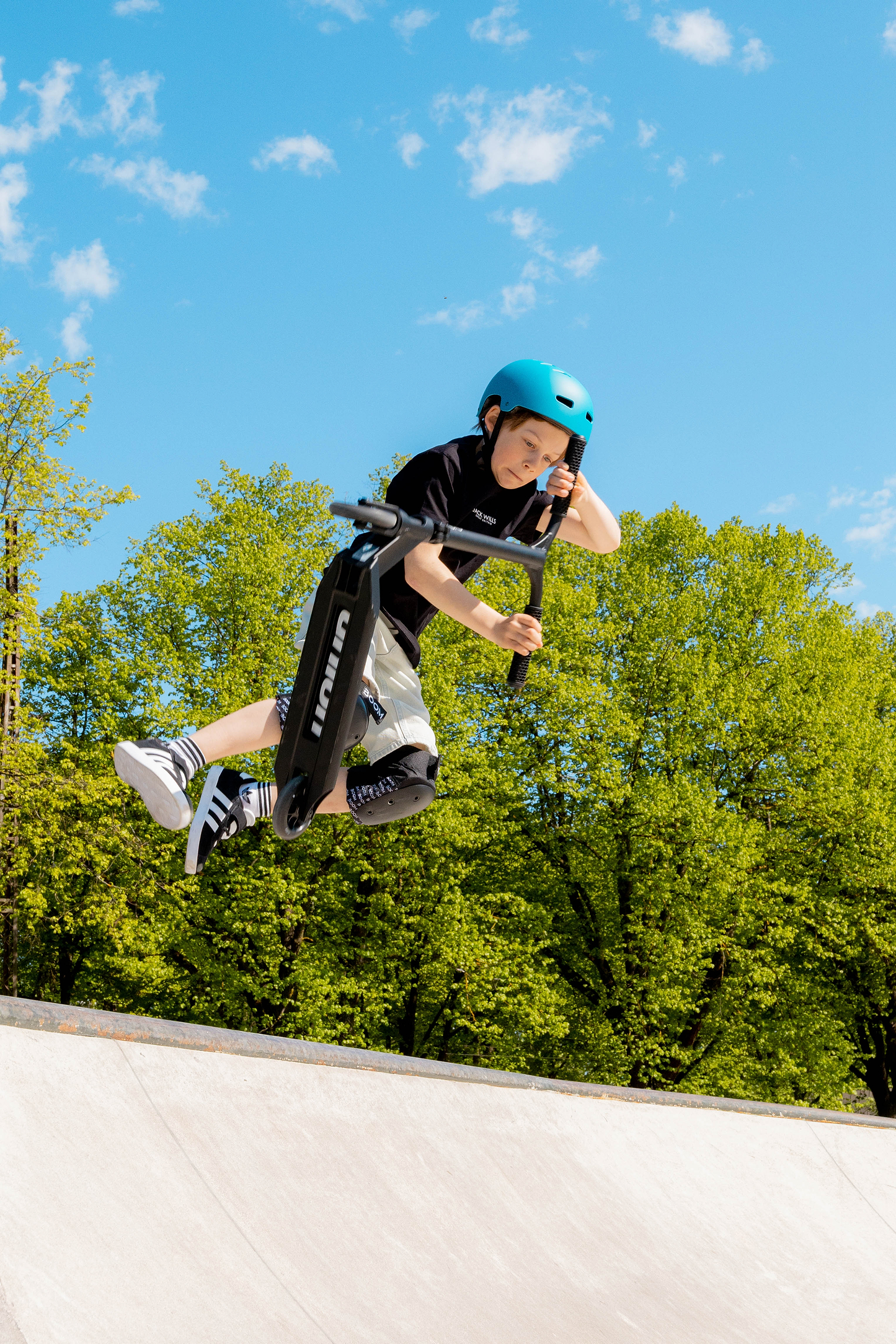A young boy wearing a blue helmet and knee pads performs a trick on the Union Aero Complete Stunt Scooter - Black by Union at a skate park, jumping above a ramp with green trees and a blue sky in the background.