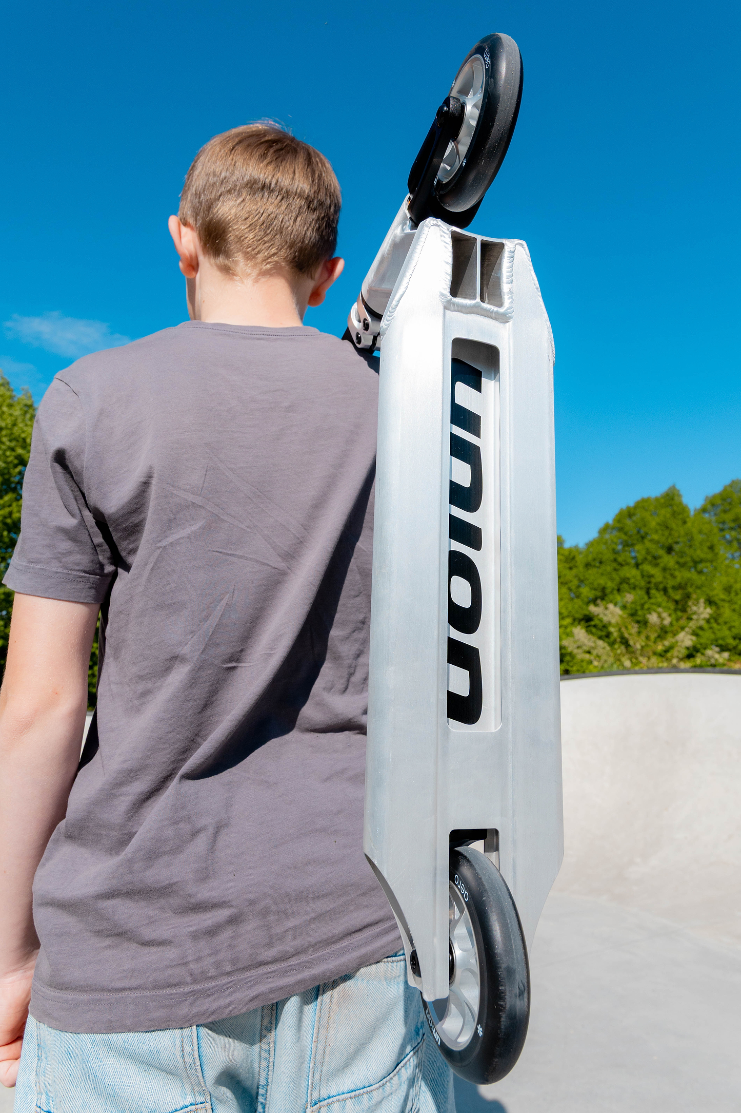 A boy in a gray shirt and light blue shorts stands outdoors, holding the Union Aero Complete Stunt Scooter - Black / Raw by Union over his shoulder. The sky is clear and blue, with green trees in the background.