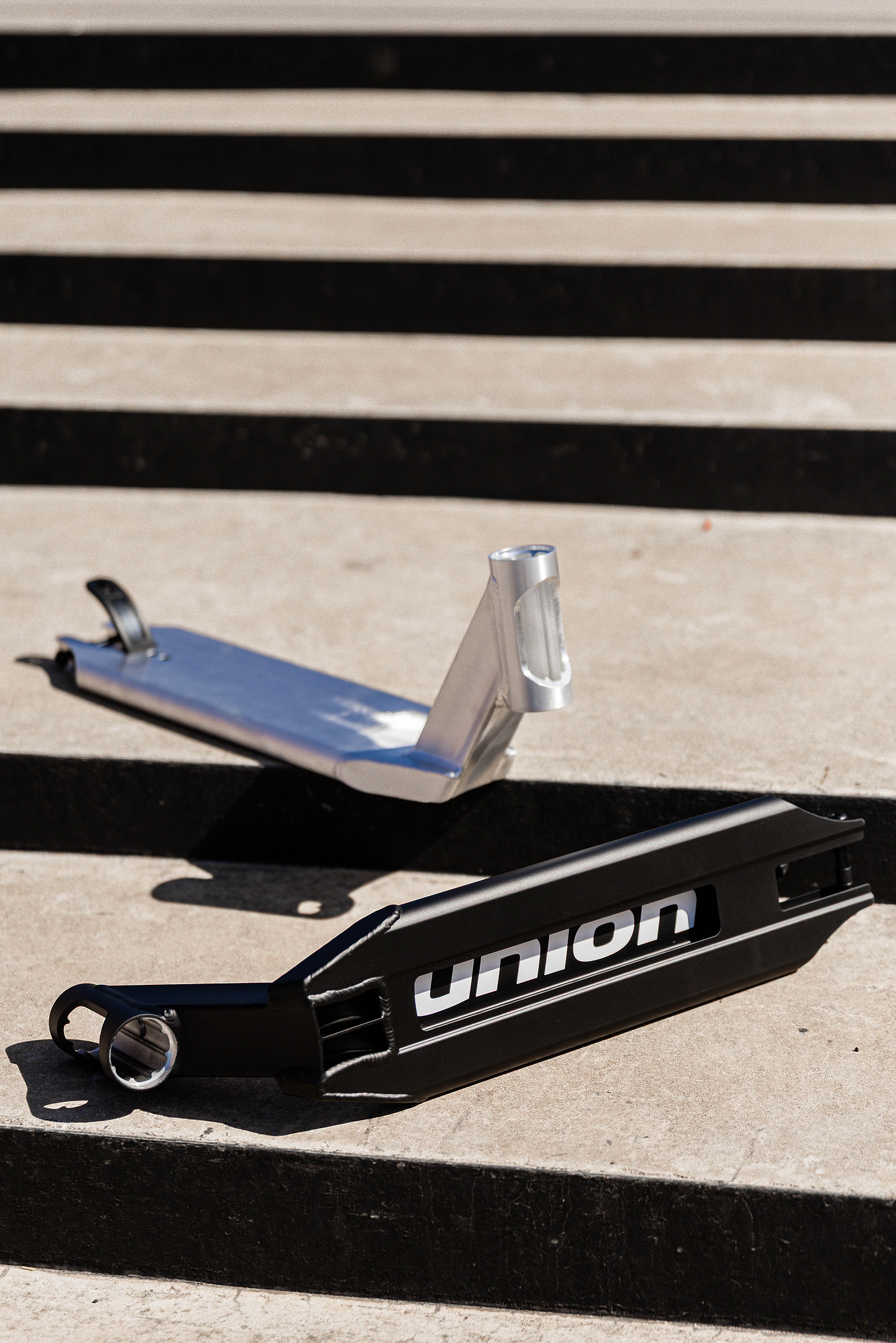 Two Union Classic Pro Silver Stunt Scooter Decks (4.75") rest parallel on sunlit concrete steps, casting sharp shadows. One deck is silver, the other black with "union" text—both from the Union brand and built lightweight for stunts.
