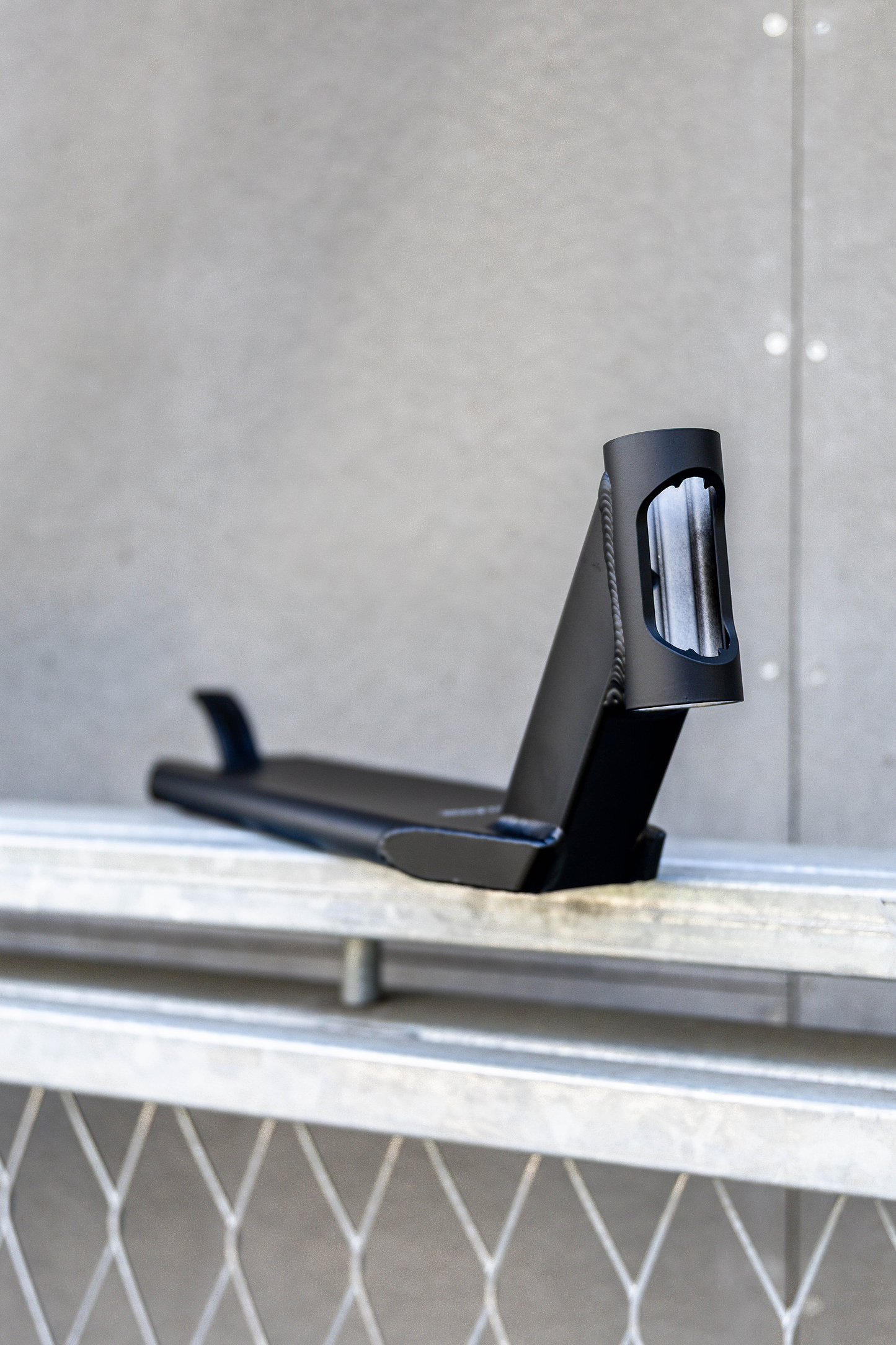 The Union Classic Pro Black Stunt Scooter Deck - 4.75" from Union rests on a metal railing in front of a gray textured wall, showcasing its smooth edges and angular design.