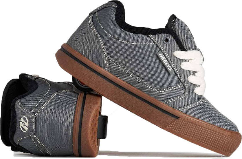 A pair of Heelys Pro 25 Boys Shoes by Heelys in Grey, Antique White, and Black is shown, with one shoe upright and the other flipped to reveal two wheels per heel. The shoes feature white laces and brown-toned soles.