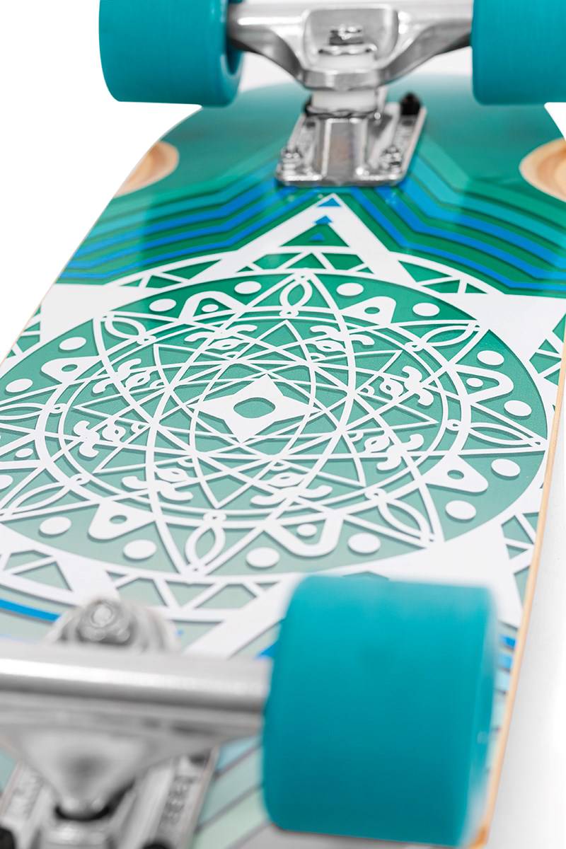 Close-up of a Mindless Drifter Cruiser Skateboard 28″ x 8″ - Teal by Mindless, showcasing teal, white, and blue geometric designs with silver trucks and blue wheels.