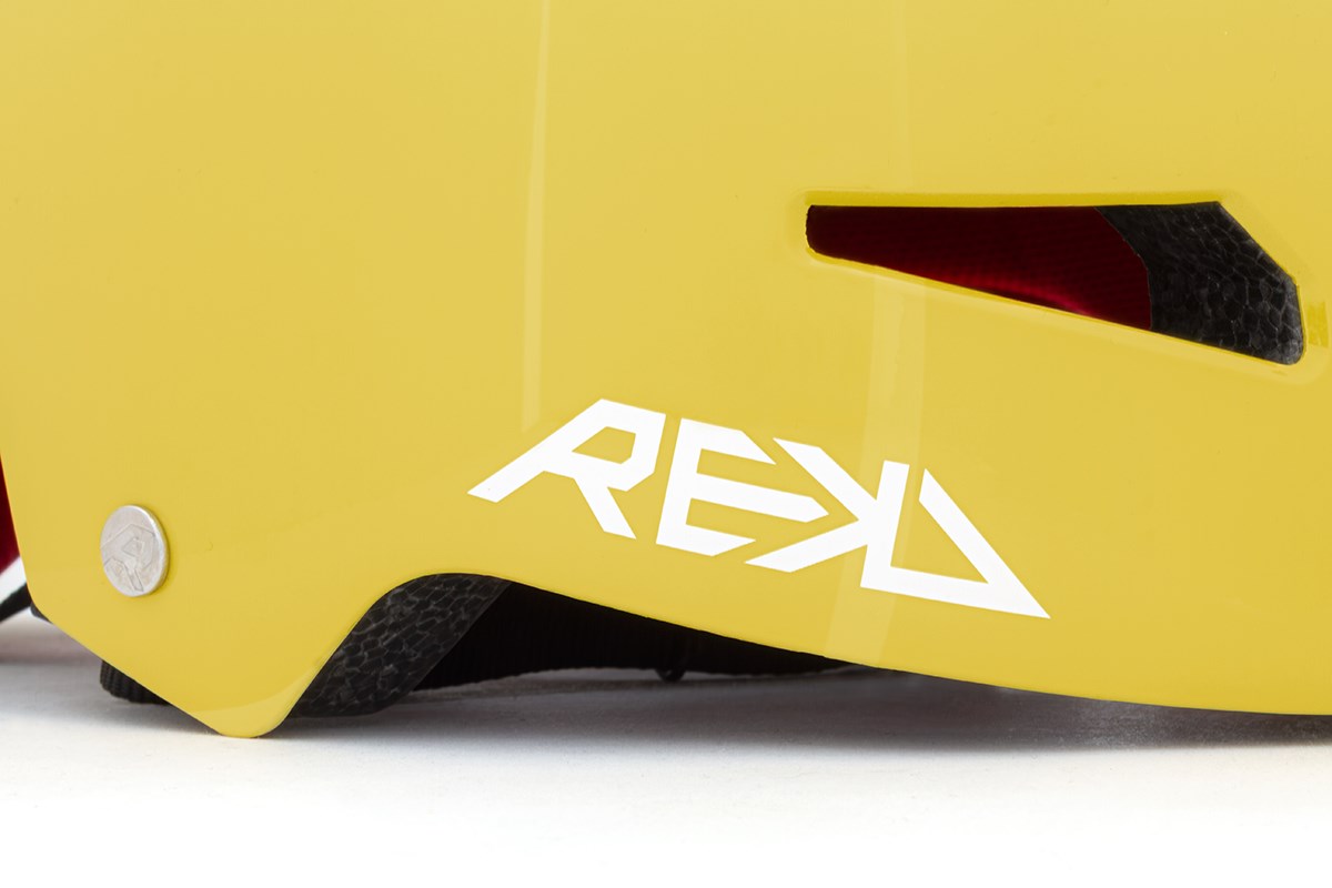 Close-up of the REKD Elite 2.0 Skate / Scooter Helmet in gloss yellow, showing the white REKD logo, small metallic rivet, partially visible black strap, and red interior padding.