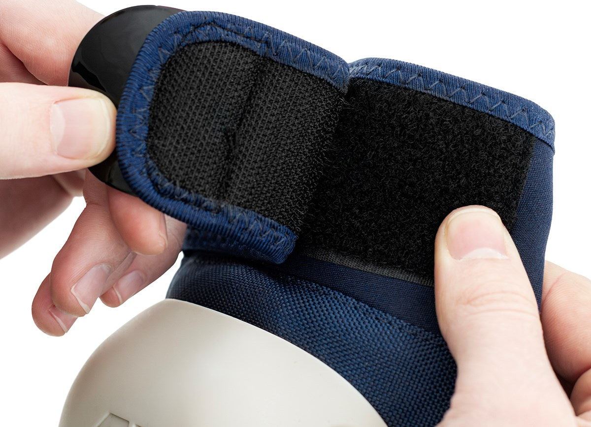 Close-up of hands fastening a black Velcro strap on the REKD Shield Triple Skate Protection Pad Set in blue, with a gray rounded section visible at the bottom.