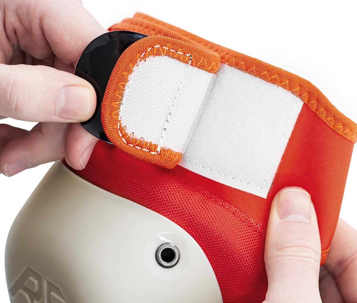 Close-up of hands fastening a white and orange Velcro strap on a REKD Shield Triple Skate Protection Pad, part of the Fade Orange / Green set designed for action sport protection on knees, elbows, or wrists.