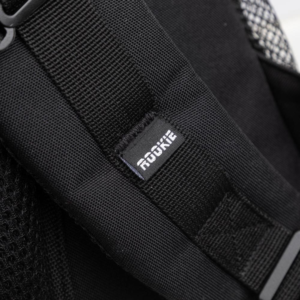 Close-up of a black fabric strap on the Rookie Skatepack Classic 78 25L Backpack by Rookie, featuring a small tag with "ROOKIE" in white text. Mesh material is partially visible on the side, showcasing the thoughtful design of this 25-liter backpack.