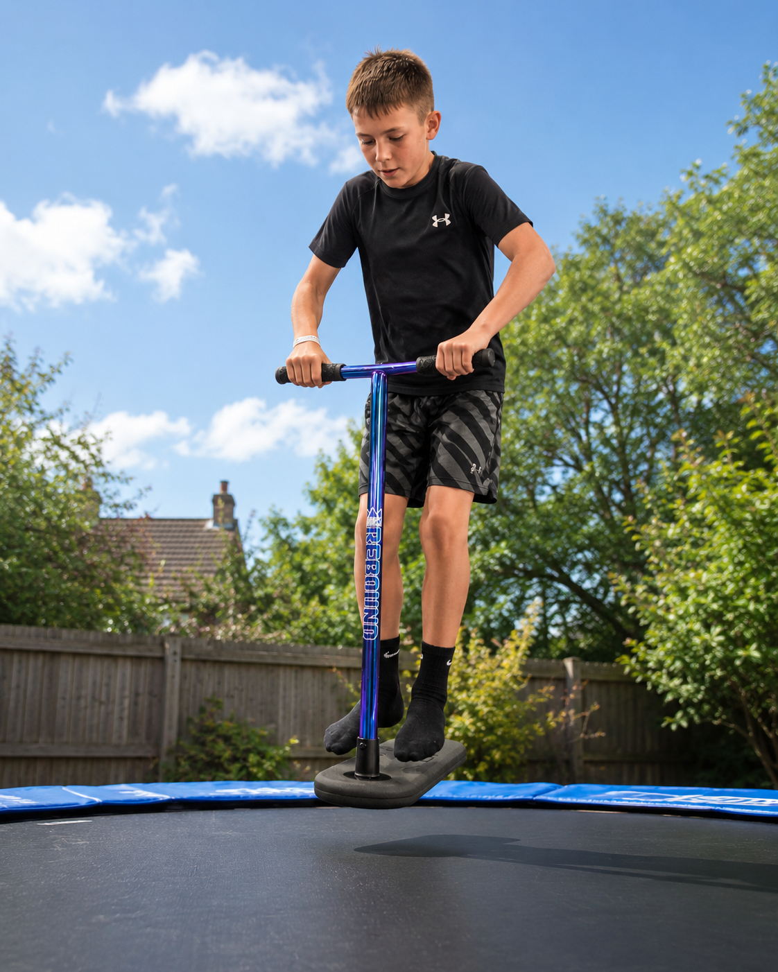 A boy in a black t-shirt and shorts jumps on a Legacy Rebound Indoor Trampoline Scooter - Blueray by Legacy on a trampoline in a backyard, surrounded by greenery, a fence, and under a partly cloudy sky.