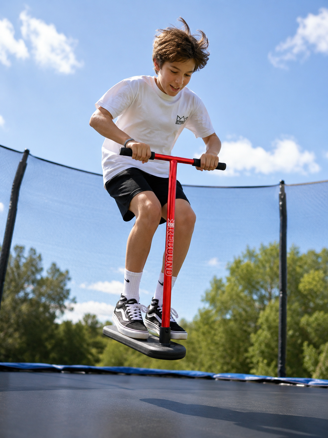 A boy in a white shirt and black shorts jumps on the Legacy Rebound Indoor Trampoline Scooter – Electro Red by Legacy. Behind him, green trees and a blue sky with clouds provide the perfect backdrop for trick training with the Legacy Rebound.