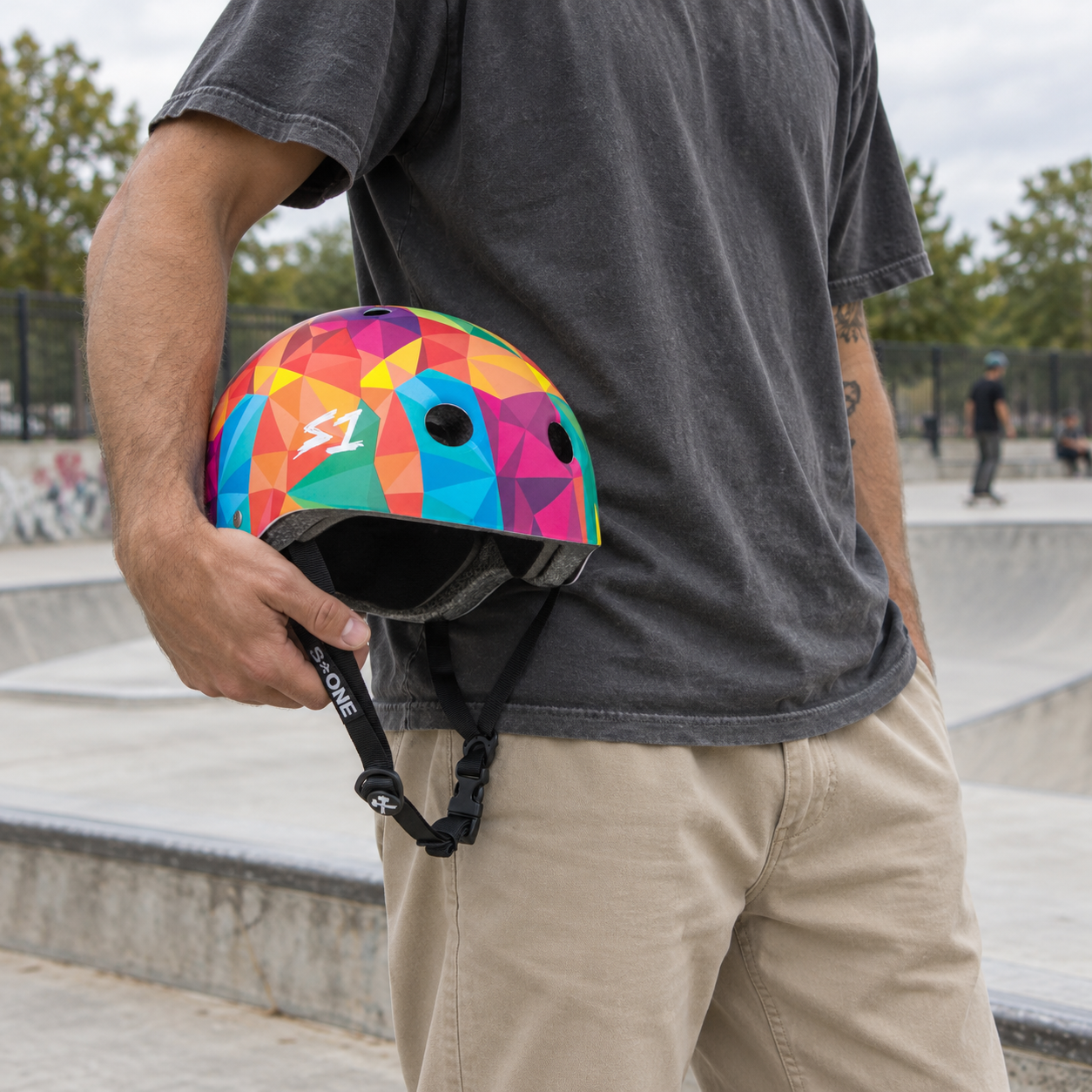 A person holding a multicolored geometric S1 Lifer Skate / Scooter Helmet - Kaleidoscope at a skatepark.