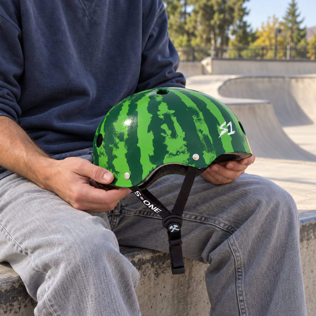 A person sits in a skatepark holding an S1 Lifer Skate / Scooter Helmet - Watermelon.