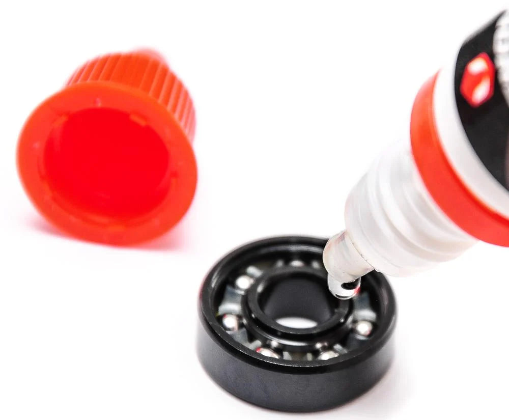A drop of Venom AntiVenom Bearing Lube 10ml Bottle lubricant falling from a nozzle onto a metal bearing, with a detached orange cap nearby, for maintaining smooth skateboard bearings.