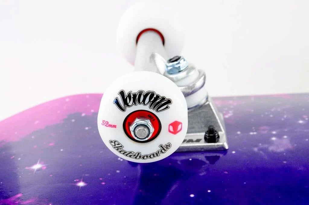 Close-up of a Venom Core Galaxy Complete Skateboard, featuring a 7.75″ x 31.75″ purple galaxy-patterned maple deck and white wheels with red and black branding.