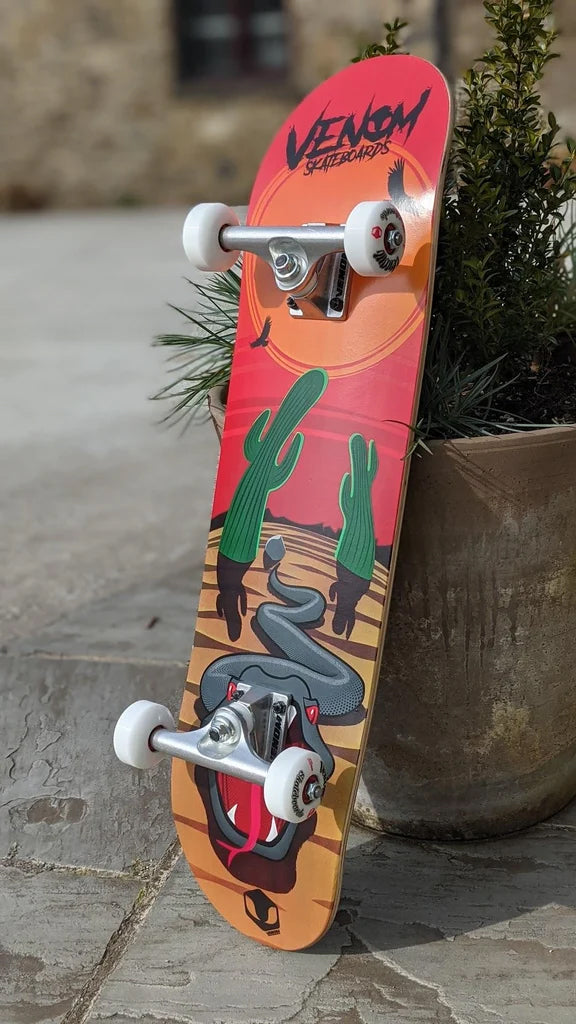 A Venom Junior Desert Viper Kids Complete Skateboard - 7.25″ x 29″ featuring a snake and cacti graphic, propped up against a terracotta pot.