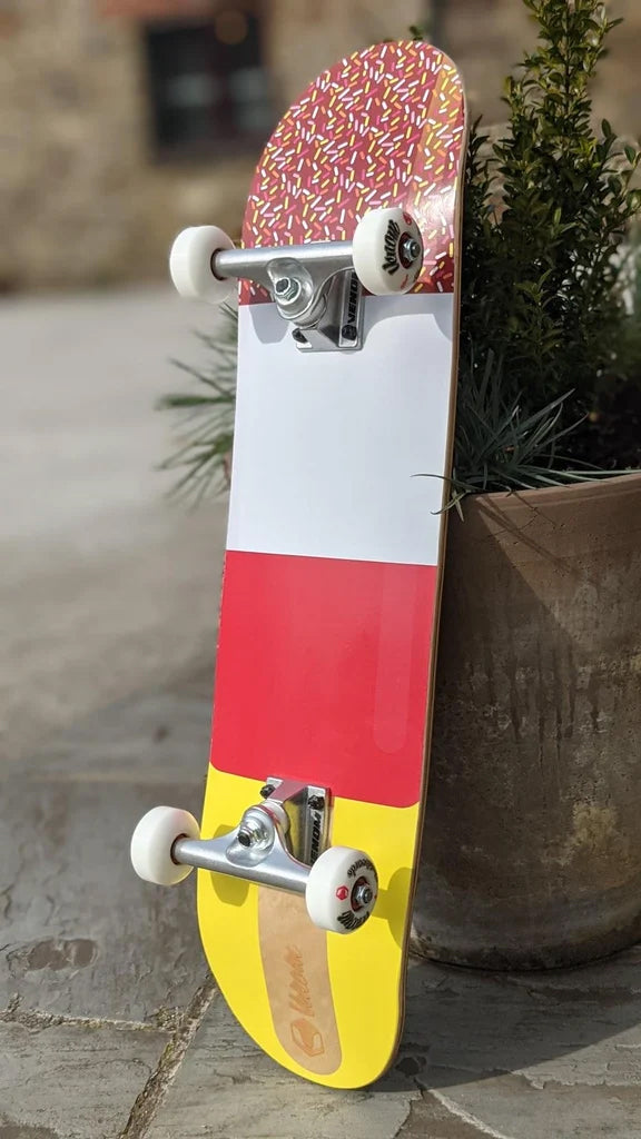 A Venom Junior Ice Lolly Kids Complete Skateboard - 7.25″ x 29″ featuring a deck with a pattern of colorful sprinkles at the top followed by white, red, and yellow sections, standing against a planter.