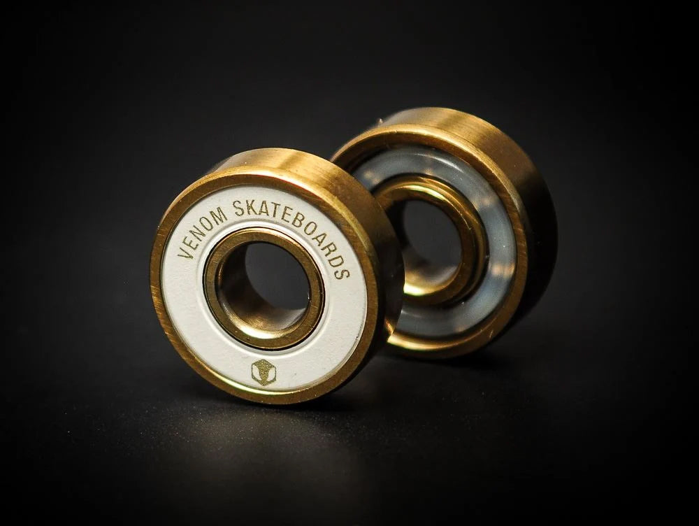 Two gold-colored Venom Precision Gold Skateboard Bearings with white central rings, each labeled with “Venom Skateboards.”