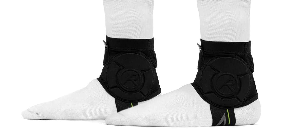 Two feet wearing white socks and black REKD Energy Covert Ankle Impact Skate Protection Guards are shown from the side. The REKD guards feature energy-absorbing circular padding and a logo, set against a plain white background.
