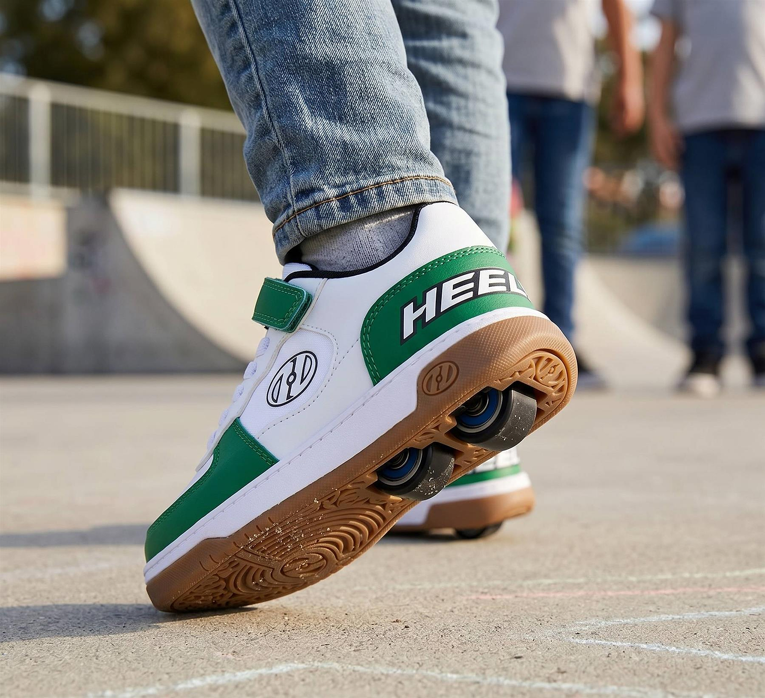 Close-up of a person wearing Heelys Rezerve Low X2 Boys Shoes in White/Green/Gum with retractable two-wheel design, standing on concrete near a skatepark. "HEEL" appears on the back of these Heelys brand shoes.