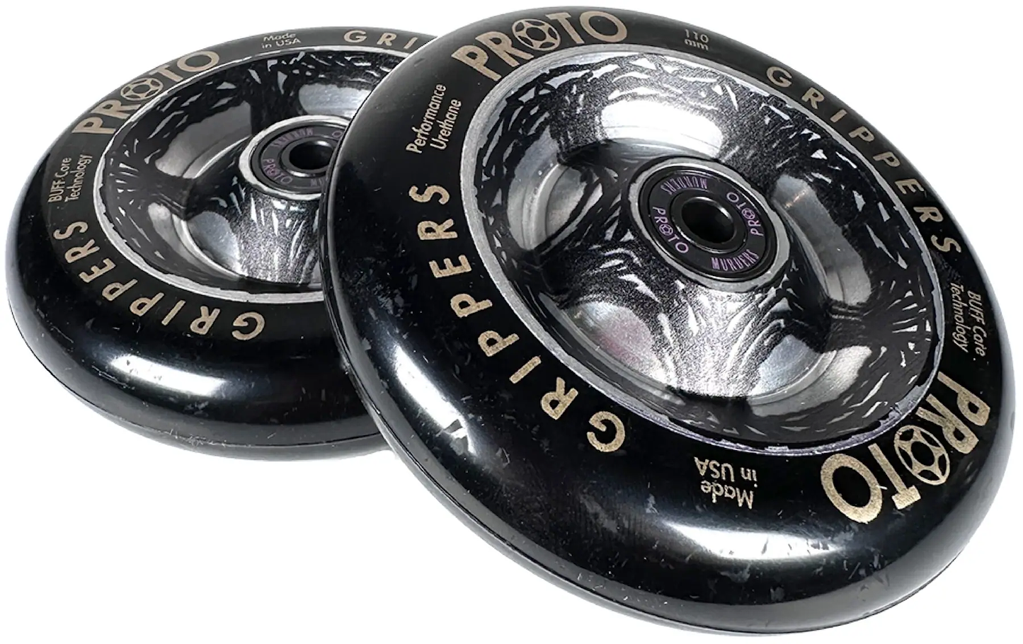 Two black Proto Catalyst Gripper 110mm stunt scooter wheels by Proto, featuring silver and gray graphics plus "PROTO GRIPPERS" and "Performance Urethane" text on the outer edge, are stacked with a slight overlap.