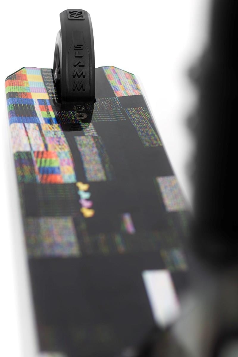 Close-up of a skateboard deck with black grip tape at the tail, displaying pixelated colorful patterns like a nebula wrap. A black wheel is at the top, suggesting agility like the sleek design of the Slamm Mischief Complete Stunt Scooter - Glitch.