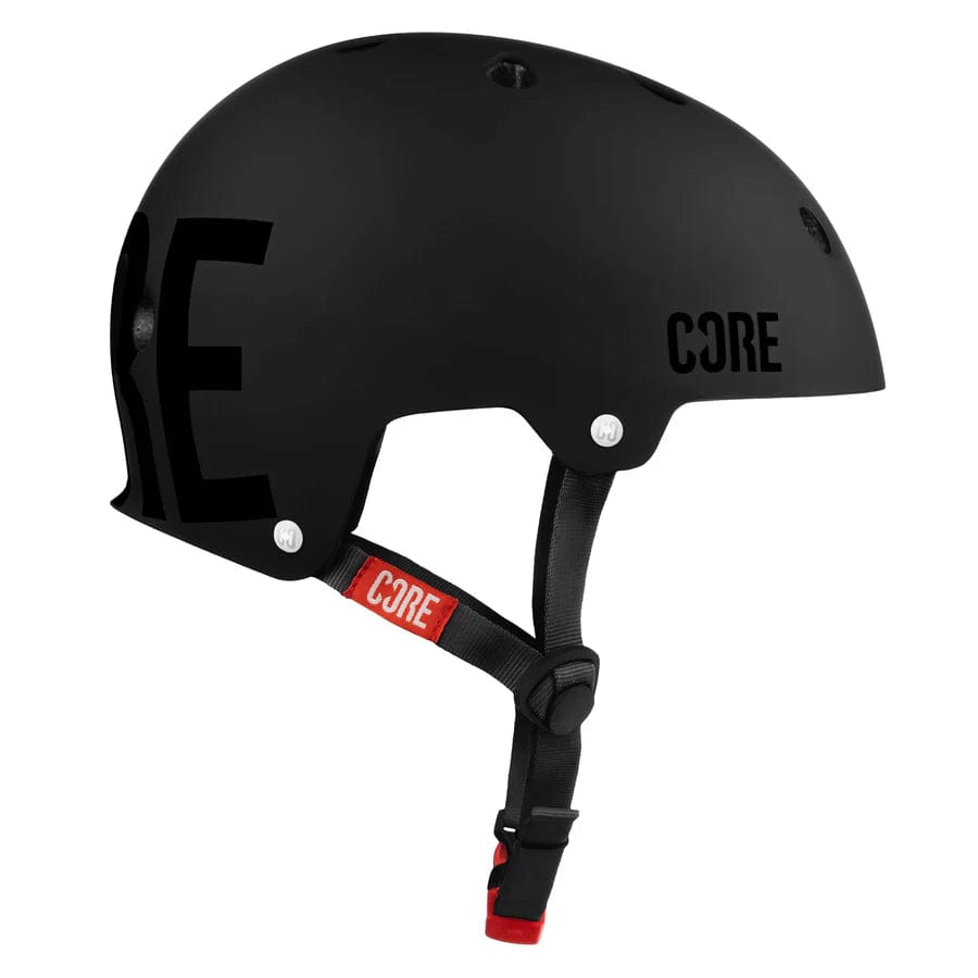 CORE Street Skate/Scooter Helmet in Stealth Black with ventilation holes and an adjustable strap. The brand name "CORE" is prominently displayed in bold black letters on the side and the strap, featuring a red accent. This low-profile helmet offers comfort while maintaining a sleek design.