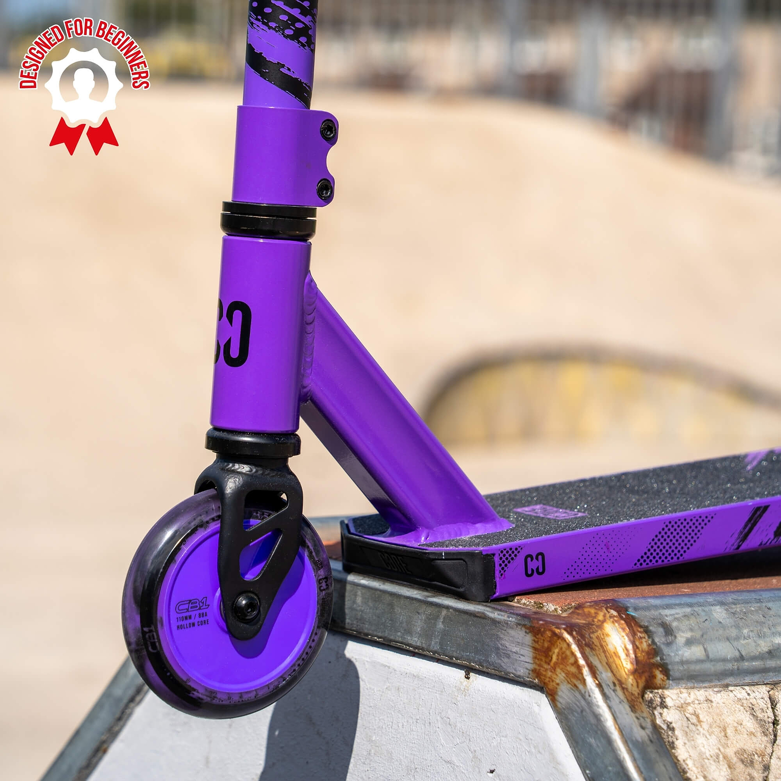 Close-up of the front wheel of a CORE CB1 Beginner Complete Stunt Scooter in Black / Purple on a skate ramp, with a “Designed for Beginners” badge in the top left corner. The scene takes place outdoors at a skatepark, highlighting this ideal choice from CORE for young riders starting out with stunt scooters.