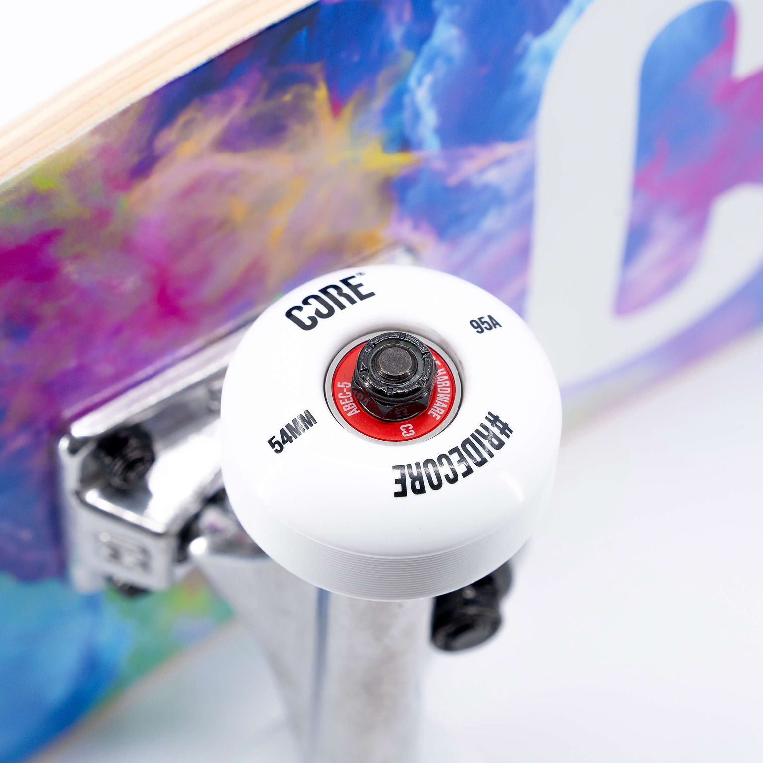 Close-up of a white 54mm skateboard wheel with a red bearing casing attached to a metal truck on a CORE C1 Neon Galaxy Complete Skateboard - 7.75″ x 31″ deck with a colorful, abstract print.