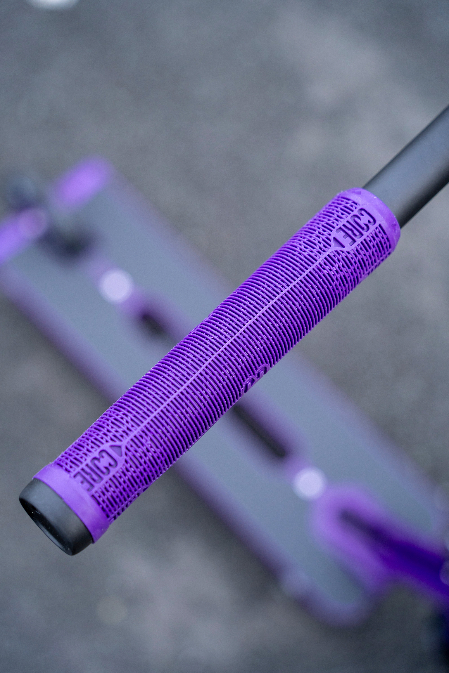 Close-up of a CORE Skinny Boy Purple Stunt Scooter Grip (170mm) on a scooter, featuring a textured pattern and the word “CORE” printed on it. The background is blurred, showing part of the scooter deck and pavement.