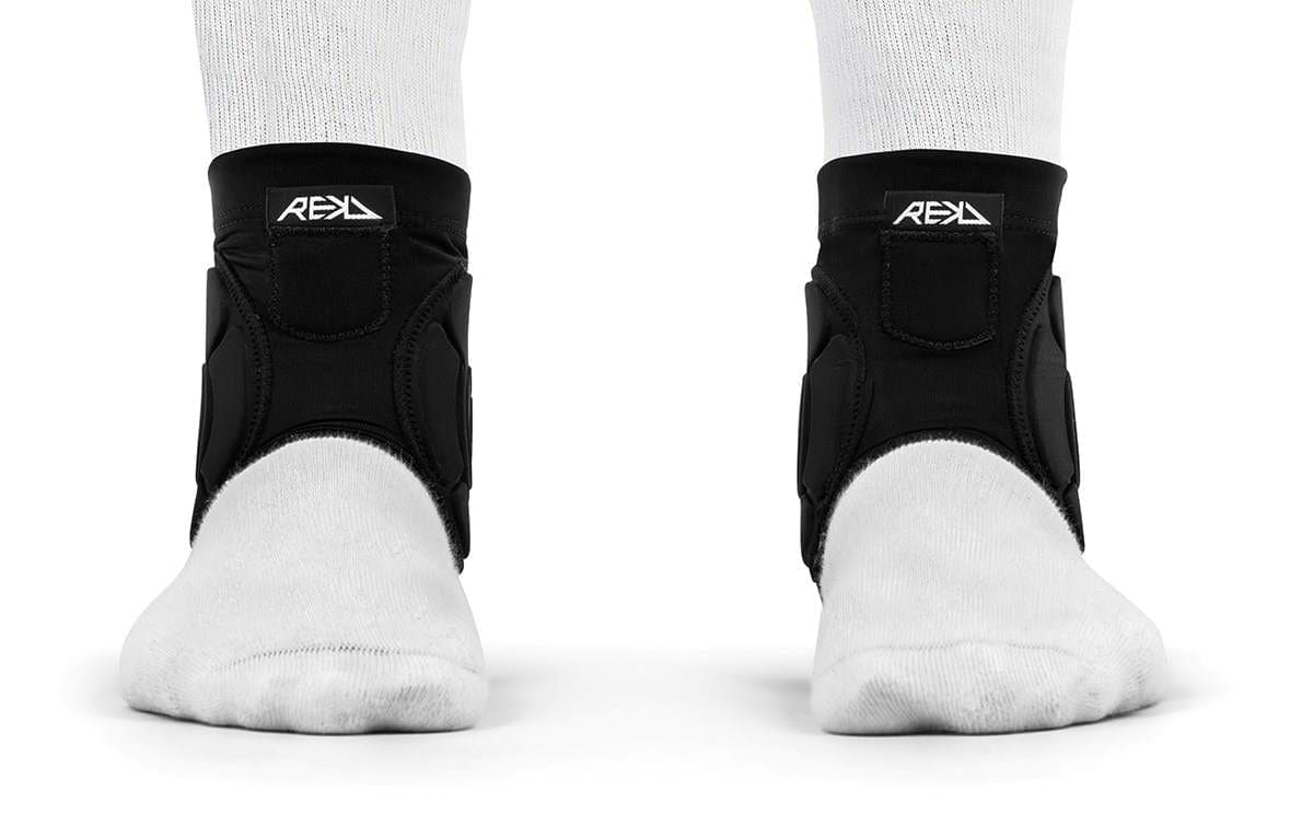 Two feet wearing white socks and black REKD Energy Covert Ankle Impact Skate Protection Guards are shown from the front on a white background. These REKD guards offer energy-absorbing protection for ankle support and stability.
