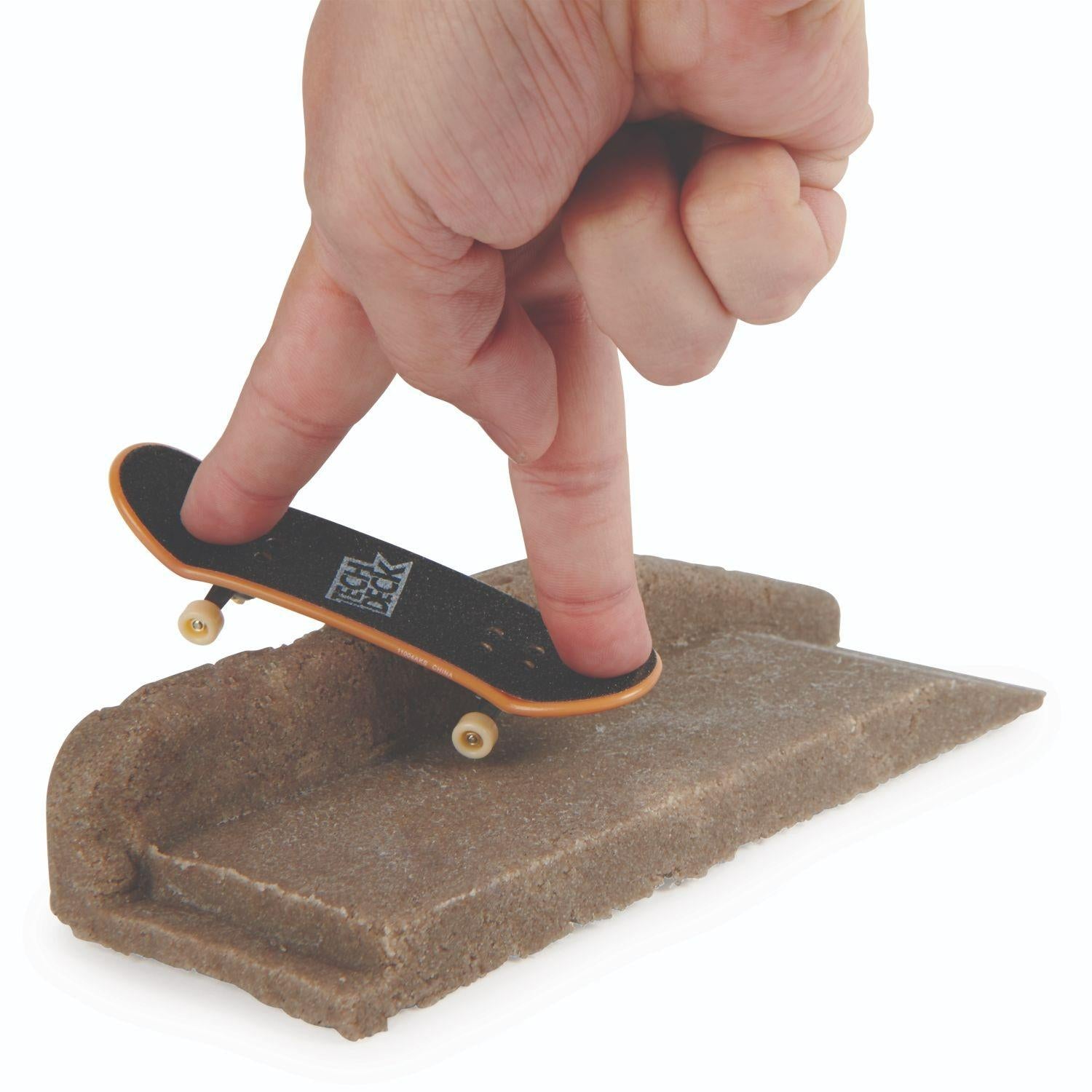 A hand uses two fingers to ride a miniature skateboard on a small brown ramp built with the Tech Deck DIY Concrete Reusable Fingerboard Ramp Modelling Set by Tech Deck, mimicking a skateboarding trick.