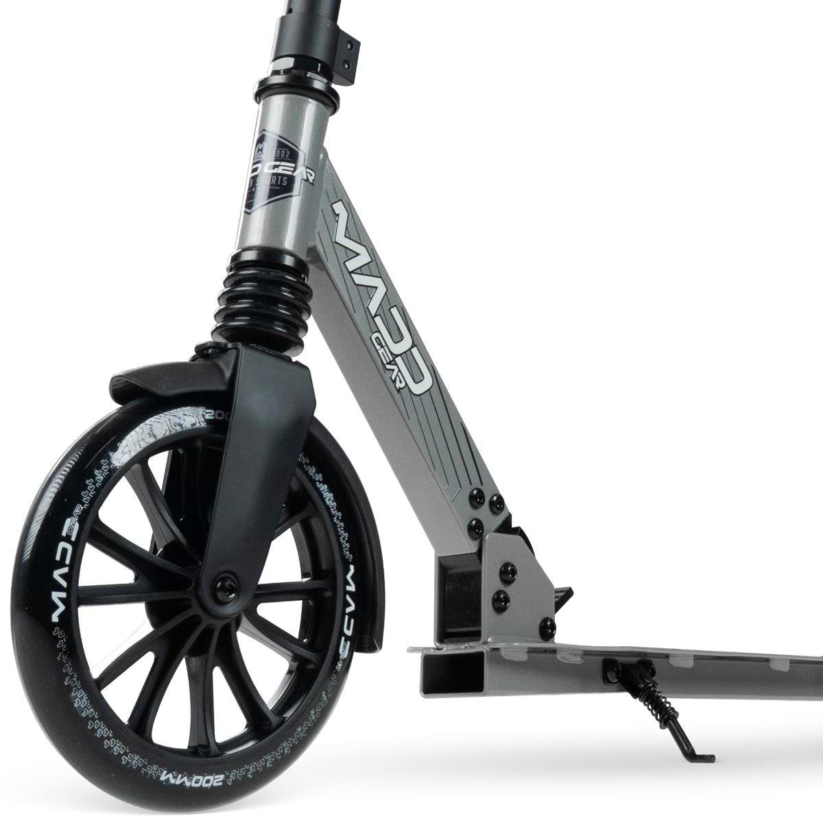 A close-up of the Madd Gear Metro Cruise 250 Foldable Commuter Scooter (Black/Grey) shows its front wheel with Madd Gear (MGP) branding, suspension fork, a portion of the deck, and a kickstand.