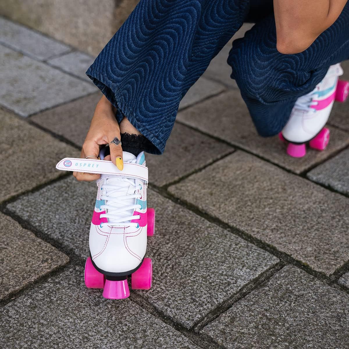 Wearing blue wavy-patterned pants, a person kneels on a stone pavement and adjusts the strap of Osprey Disco Quad Roller Skates - White with bright pink wheels, ready for fun in these stylish Osprey recreational skates.