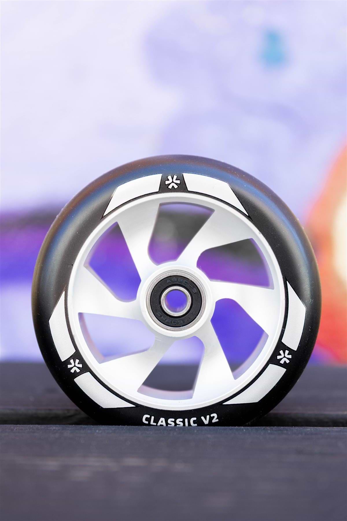 A close-up of the Union Classic V2 110mm Stunt Scooter Wheel in Black/Silver by Union, featuring "CLASSIC V2" text, upright on a dark surface with a blurred, colorful background.