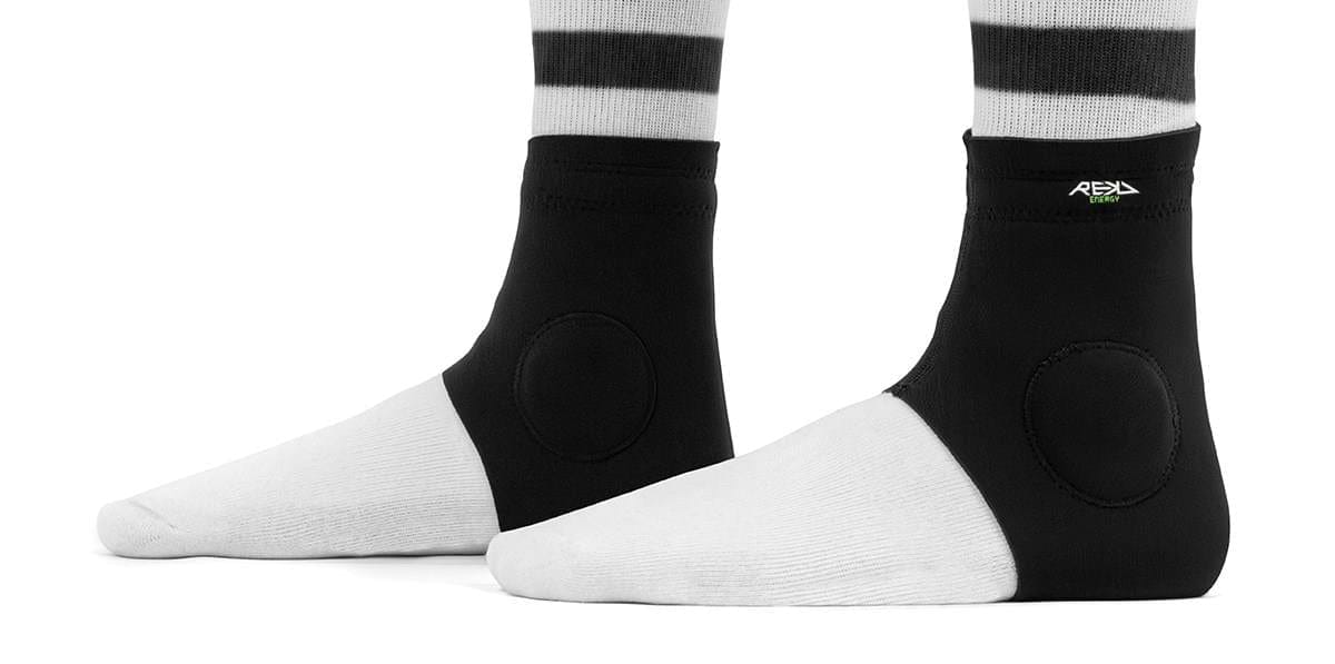 Two feet in white socks wear REKD Energy Slim Footies Ankle Protection Pad - Black by REKD, featuring circular side padding and a small green-and-white REKD Energy logo near the top. The background is plain white.