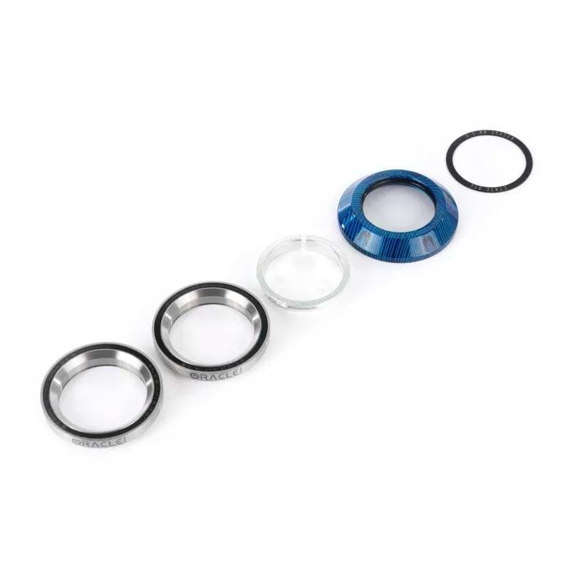 The Ethic DTC Oracle Integrated Stunt Scooter Headset in Blue is presented with its disassembled mechanical components aligned horizontally. It showcases a circular metal ring in black, silver, and blue shades, reminiscent of a stunt scooter headset. Additionally, it includes a clear glass lens and a thin black gasket designed to reduce friction and enhance compression.