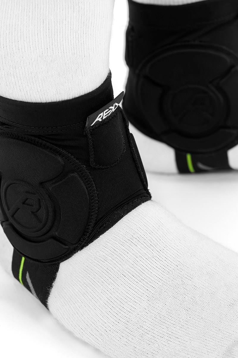 Close-up of a person wearing white socks and black REKD Energy Covert Ankle Impact Skate Protection Guards, featuring the REKD brand label. These guards deliver energy-absorbing protection to help reduce impact forces.