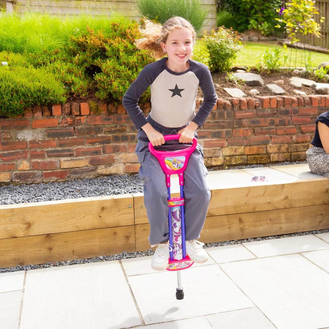 A smiling girl with curly hair bounces outdoors on an Ozbozz Unicorn Kids Pogo Stick - Pink, wearing a star shirt and gray pants, with greenery and a brick wall behind her.