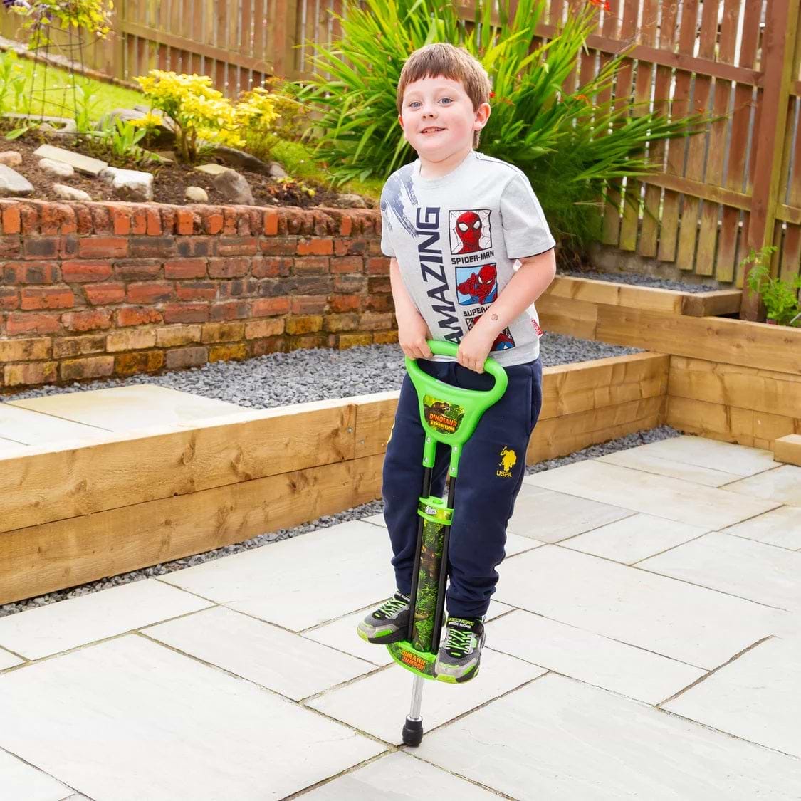A young boy in a Spider-Man T-shirt and navy pants smiles while jumping on the Ozbozz Dinosaur Kids Pogo Stick - Green by Ozbozz, on a backyard patio with stone tiles and wooden fences.
