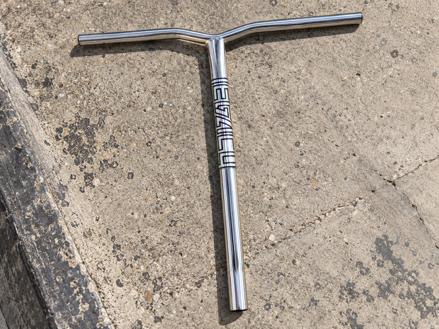 Legacy Ventus Steel Standard IHC Stunt Scooter Bars in chrome featuring a T-style design and a vertical graphic on the down tube, laid on a concrete surface.