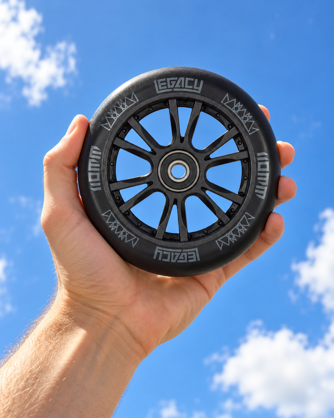A hand holds a black Legacy Ventus 110mm Stunt Scooter Wheel against a blue, cloudy sky.