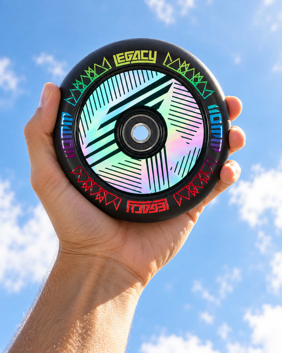 A hand holds a black 110mm Legacy Ventus 1.5 Pro 110mm Stunt Scooter Wheel - Neochrome with a holographic, geometric hollow core design against a blue, cloudy sky.